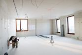 A person wearing safety equipment aligns wall markings using a laser leveling tool in a bright room.